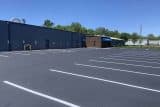 A large, empty parking lot with freshly done asphalt paving and marked spaces leads to a blue and brick building in the distance. The sky is clear and blue, with trees along the horizon. An American flag is visible on the right side.