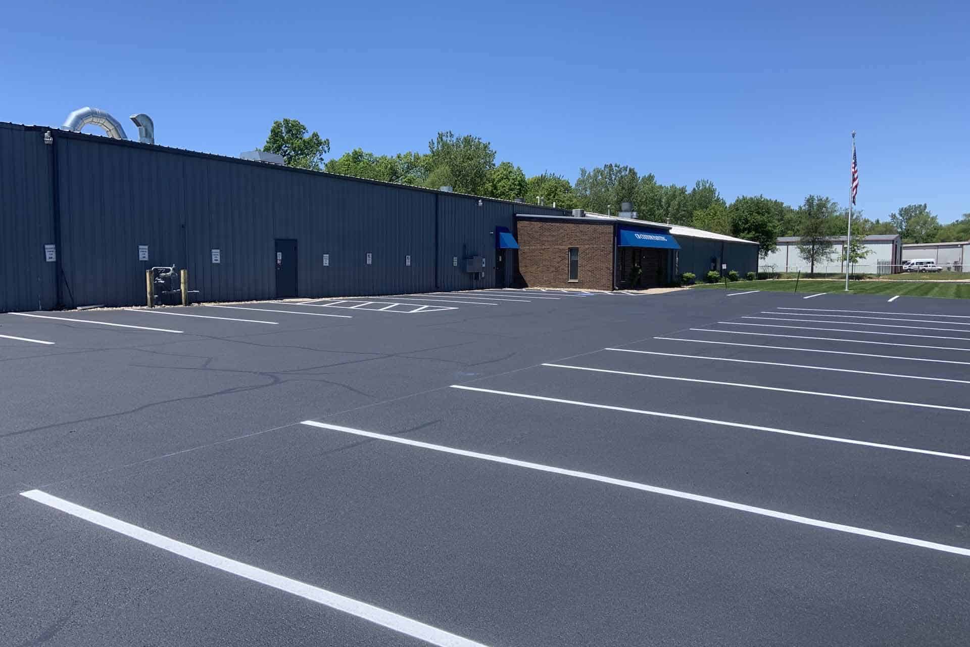 A large, empty parking lot with freshly done asphalt paving and marked spaces leads to a blue and brick building in the distance. The sky is clear and blue, with trees along the horizon. An American flag is visible on the right side.